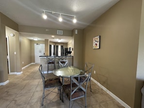 A dining area with a round glass table and metal chairs, a kitchen in the background, and a hallway leading to a door. - 2306 Spacious 2/2 Resort Condo Near Disney (Orlando)