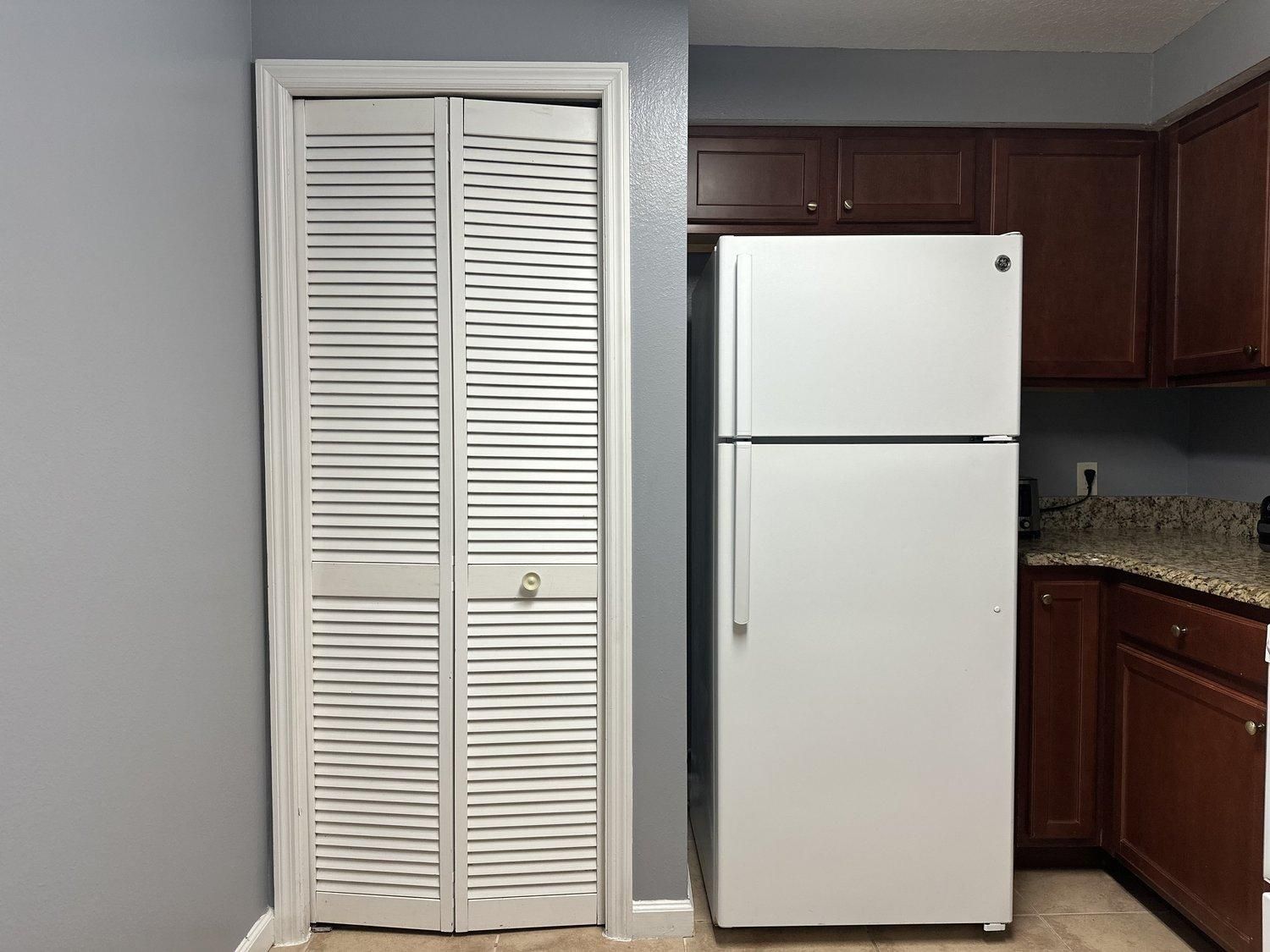 Une cuisine avec un réfrigérateur blanc, des armoires en bois et une porte blanche fermée dotée de stores horizontaux.