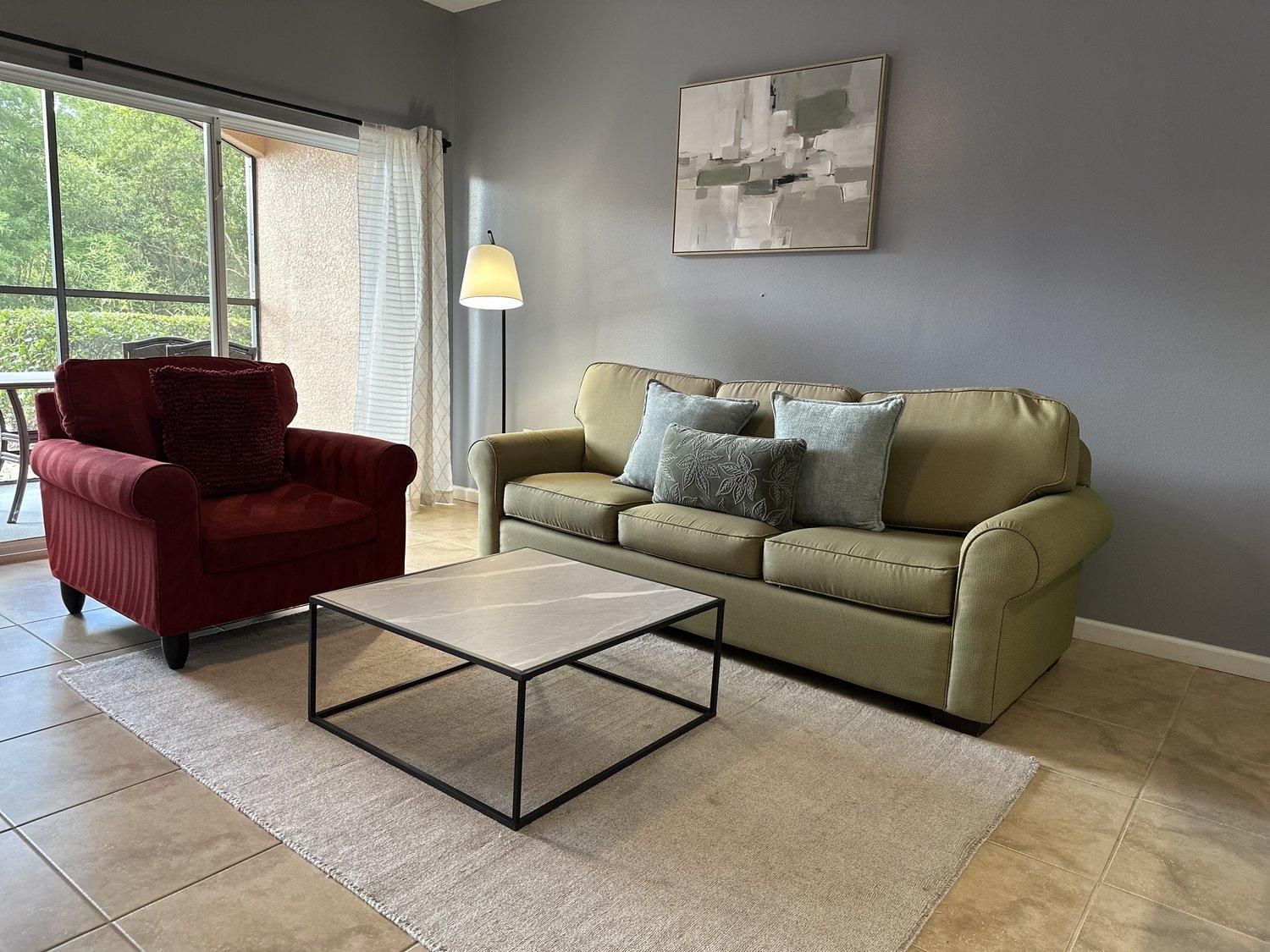 Un salon avec un fauteuil rouge, un canapé vert orné de coussins à motifs, une table basse et un lampadaire.