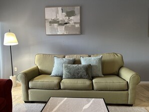 Ein Wohnzimmer mit einem grünen Sofa, zwei gemusterten Kissen, einem Marmortisch, einer Stehlampe und einem abstrakten Kunstwerk an der Wand.