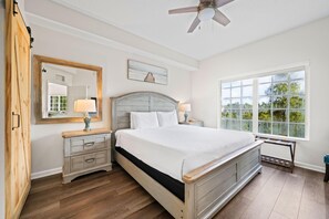Ein Schlafzimmer mit Bett, Nachttischen, einem Schminktisch und einem Deckenventilator, mit Blick auf Bäume.