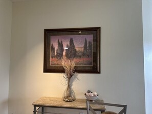 Ein gerahmtes Landschaftsbild an der Wand, eine Vase mit Trockenpflanzen auf einem Tisch und eine dekorative Schale.