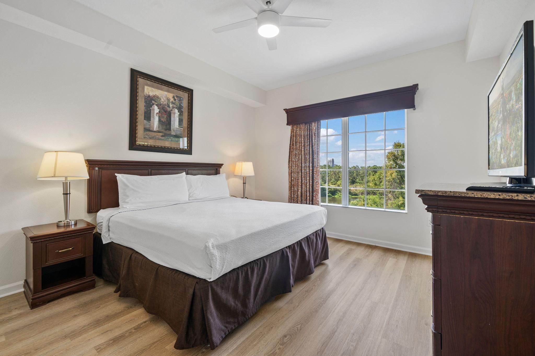 Une chambre d’hôtel avec un grand lit, deux lampes de chevet, un ventilateur de plafond, une fenêtre avec des rideaux et un tableau au mur.
