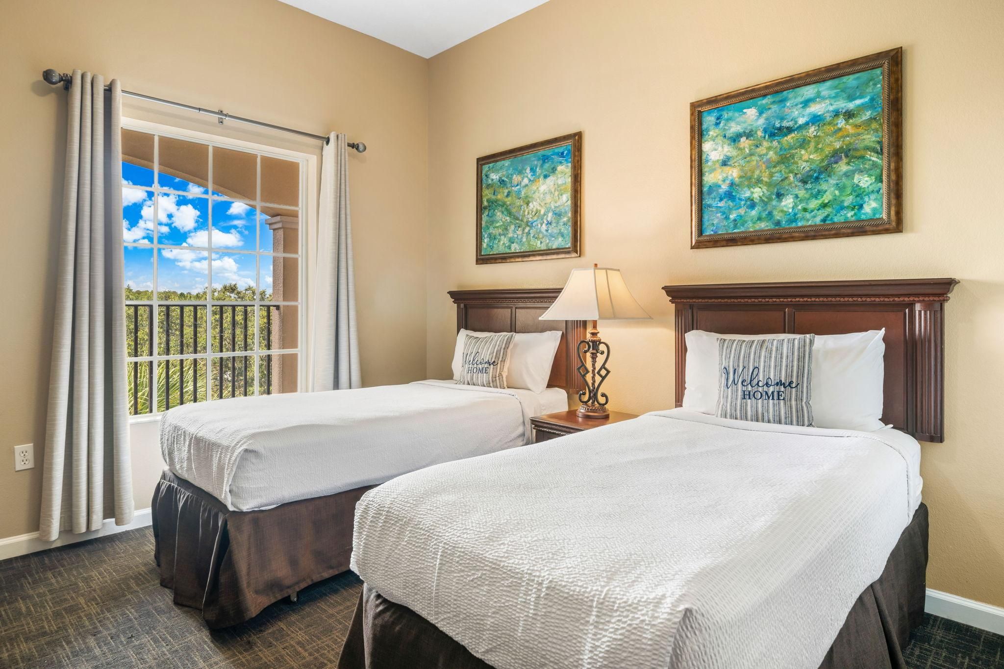 Une chambre d’hôtel avec deux lits, une fenêtre avec des rideaux, une lampe et deux tableaux au mur.