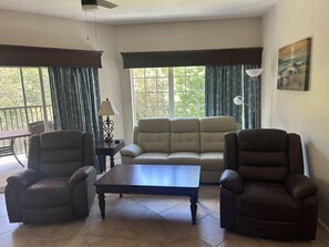 Ein Wohnzimmer mit einer Couch, zwei Sesseln, einem Couchtisch und einer Lampe. Große Fenster mit Vorhängen.