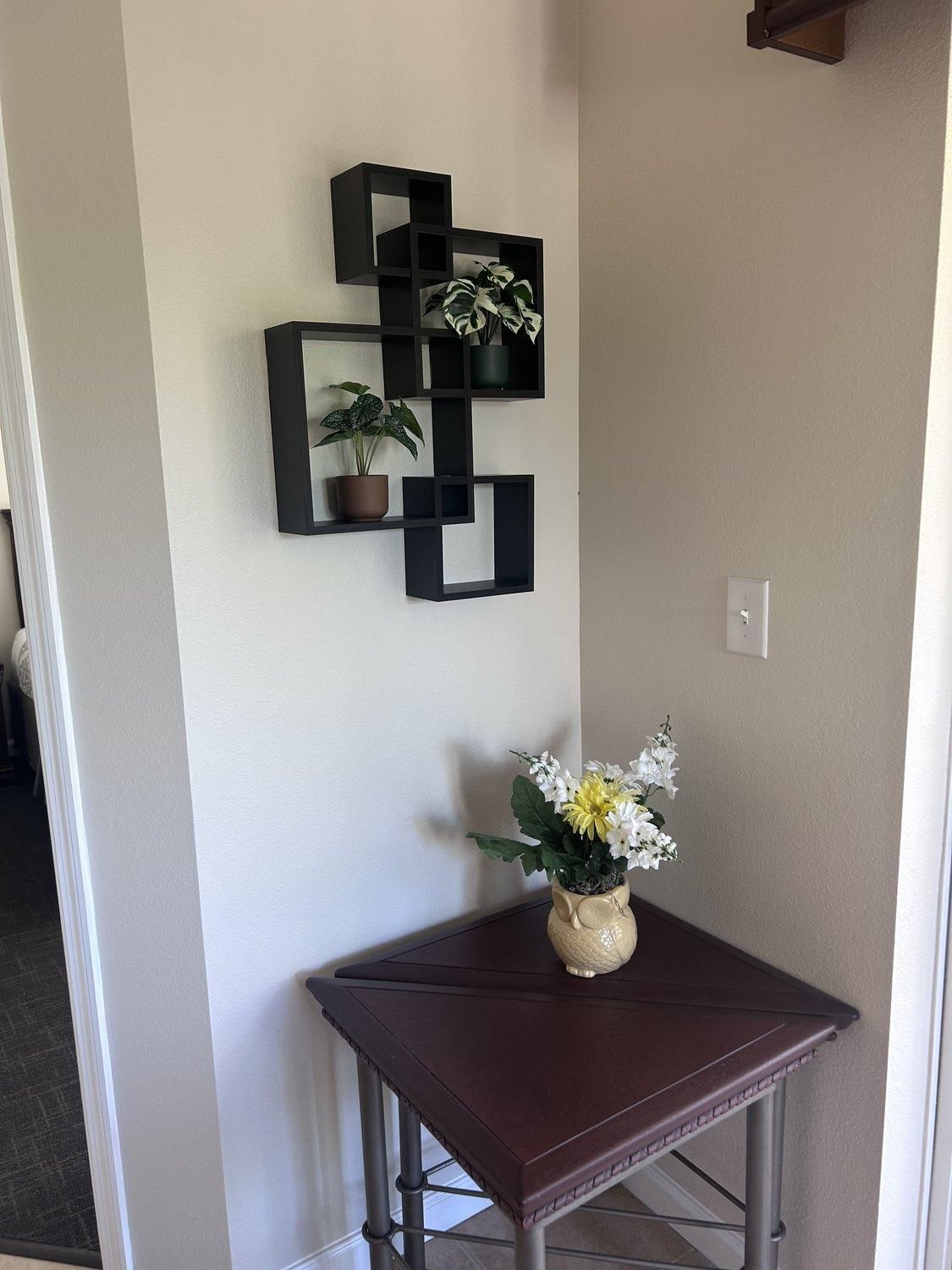 Un coin avec une table marron foncé, une étagère noire fixée au mur où sont posées des plantes en pot, et un vase rempli de fleurs.