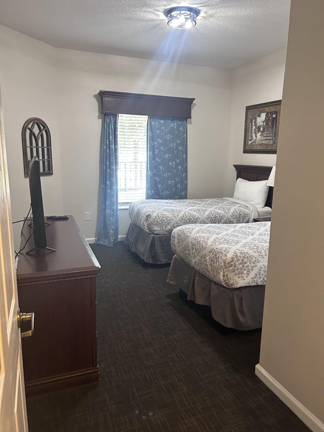 Une chambre d’hôtel avec deux lits, un bureau, une fenêtre avec des rideaux et un luminaire au plafond.