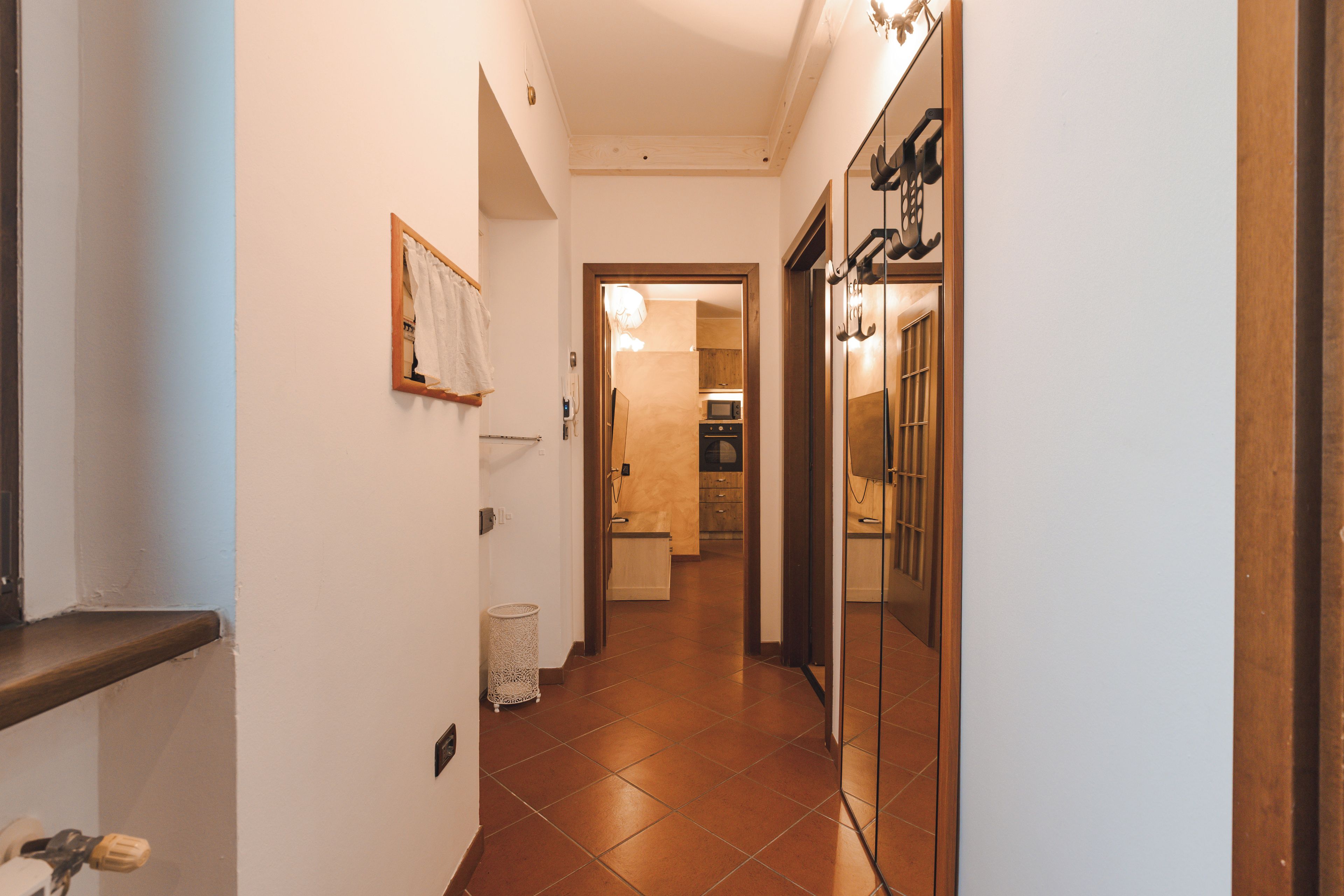 A narrow hallway with a wooden floor, a white wall, and a door leading to a kitchen area.
