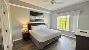 Ein Schlafzimmer mit Bett, Nachttischen, Deckenventilator, einem Fenster mit Vorhängen und einem Bild an der Wand.