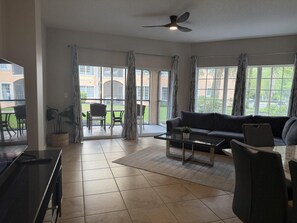 Una sala de estar con un sofá, una mesa de centro y un televisor. Hay cortinas en las ventanas y se ve un ventilador de techo.
