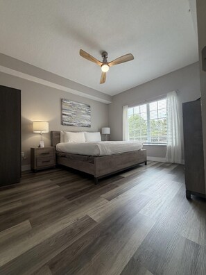 A bedroom with a bed, bedside tables, a ceiling fan, a window with curtains, and a painting on the wall.