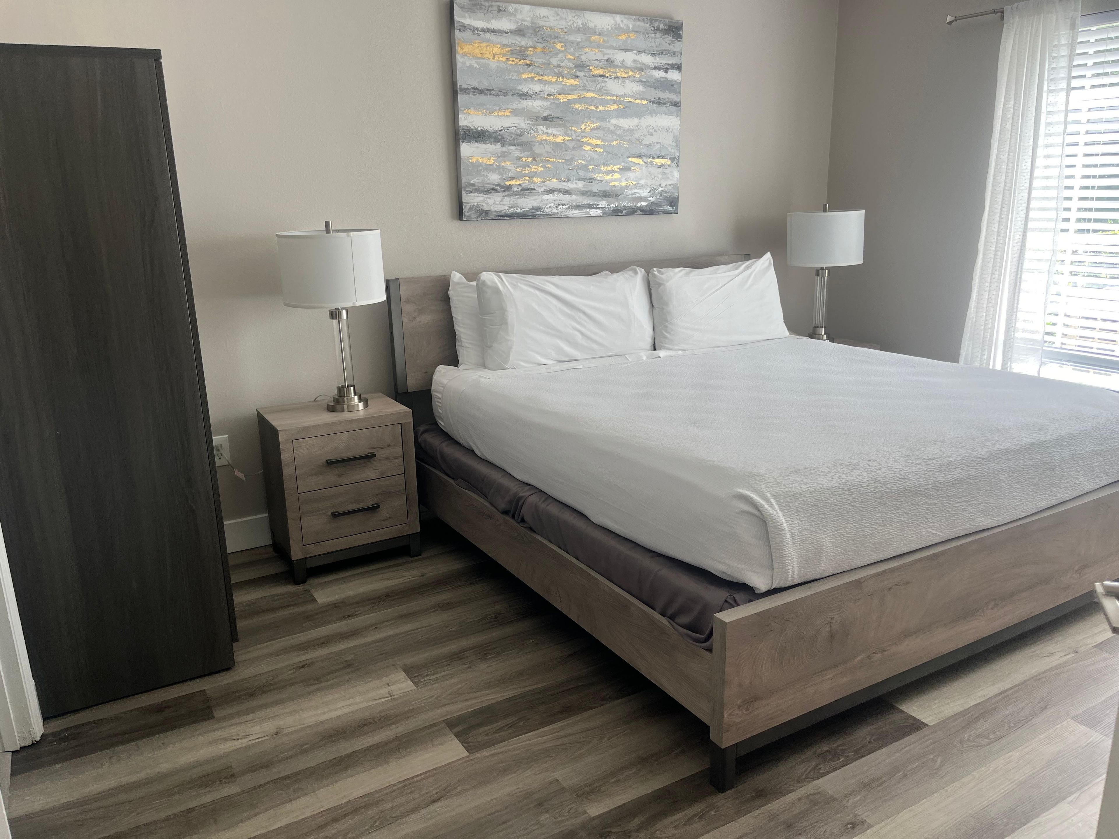 Une chambre à coucher comprenant un lit, des tables de chevet, une commode, un tableau accroché au mur et une fenêtre avec des stores.