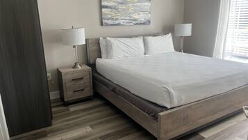 Une chambre à coucher comprenant un lit, des tables de chevet, une commode, un tableau accroché au mur et une fenêtre avec des stores.