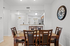 Un comedor con mesa y sillas de madera, una cocina al fondo y un reloj de pared.