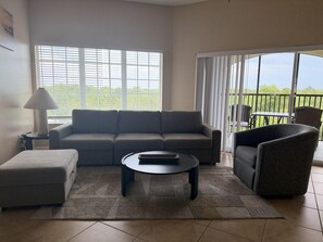 Ein Wohnzimmer mit einer Couch, einem Hocker, einem Couchtisch und einem Sessel. Ein Fenster mit Jalousien, eine Lampe und Blick ins Grüne sind zu sehen.