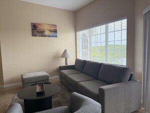 Ein Wohnzimmer mit einer Couch, einem kleinen Tisch und einer Lampe. An der Wand hängt ein Gemälde und es gibt ein Fenster mit Jalousien.