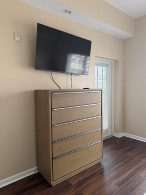 Ein Zimmer mit einem an der Wand befestigten Flachbildfernseher, einer Holzkabine mit Schubladen und einem Fenster mit Jalousien.