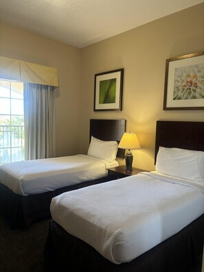 Habitación de hotel con dos camas, una mesita de noche con lámpara, una ventana con cortinas y dos cuadros en la pared.
