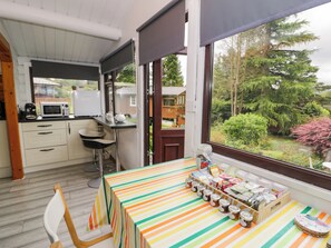 A room with a kitchen area, a dining table with striped tablecloth, and a view of a garden. - Babbling Brook (Blaenau Ffestiniog)