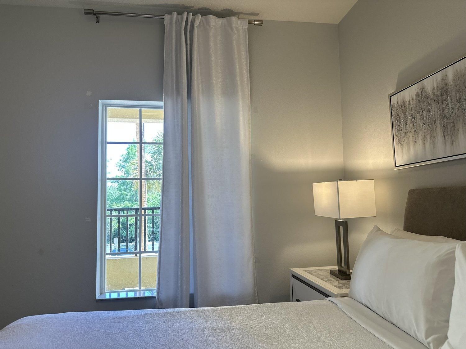 Une chambre d’hôtel avec un lit, une table de chevet avec une lampe, une fenêtre avec des rideaux et un tableau au mur.