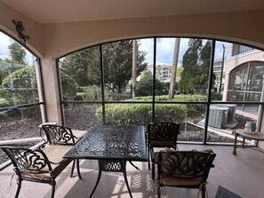 Um alpendre coberto com mesa e cadeiras, grandes janelas com vista para uma área ajardinada e edifícios.