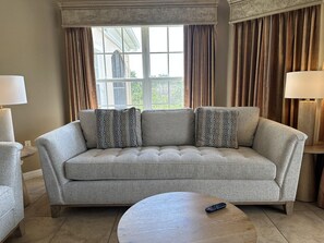 A living room with a grey sofa, two patterned throw pillows, a round wooden coffee table, and a remote control on it. - 1207 Spacious 3/2 Resort Condo Near Disney (Orlando)