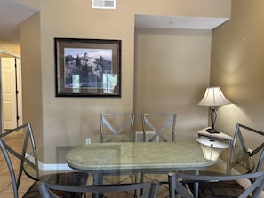 A dining area with a glass table and metal chairs, a framed picture on the wall, and a lamp on a side table. - 1207 Spacious 3/2 Resort Condo Near Disney (Orlando)