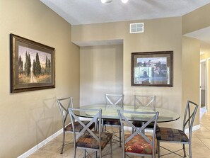 A dining area with a glass table and metal chairs, framed pictures on the walls, and a ceiling vent. - 1302 Spacious 3/2 Resort Condo Near Disney (Orlando)