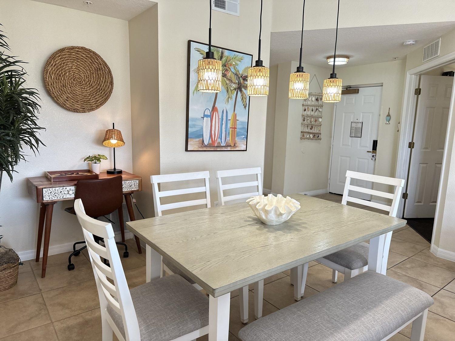 A dining area with a light-colored table, white chairs, a woven placemat, a framed painting, and pendant lighting.