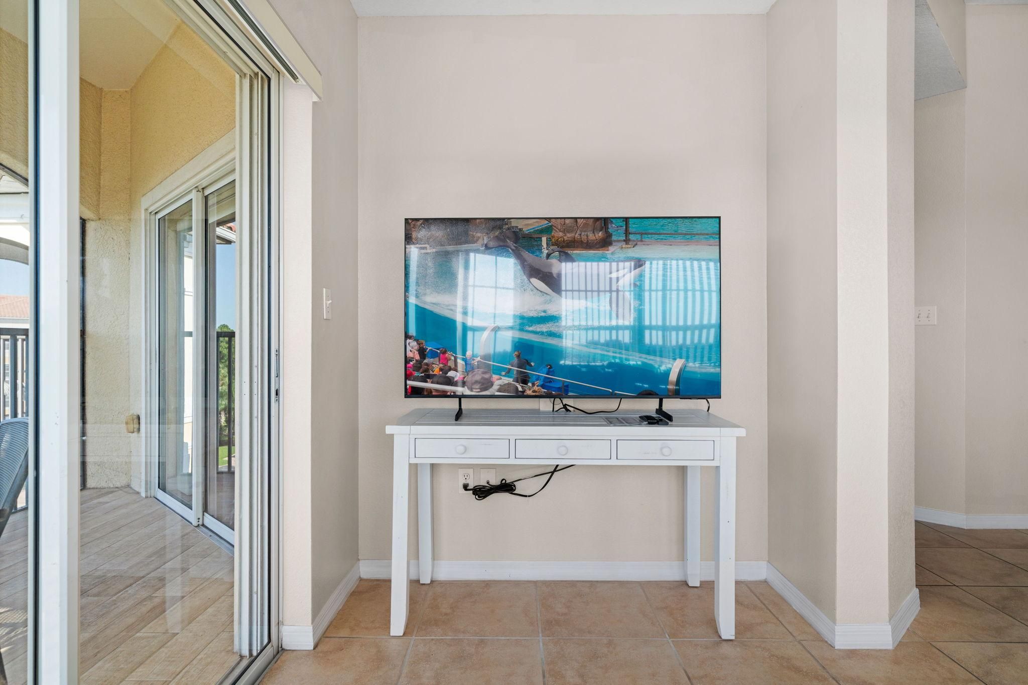 Una habitación con una televisión instalada en una mesa consola blanca, un ventanal corredero que da a un espacio exterior y suelo de baldosas.