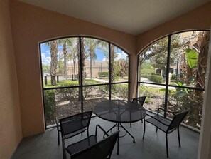 A room with a round table and four chairs, large windows with a view of palm trees and a building, and a concrete floor. - 2101 Spacious 3/2 Resort Condo Near Disney (Orlando)