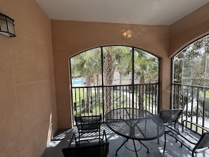 A sunlit balcony with a round metal table and four chairs, overlooking a pool and palm trees. - 2201 Spacious 3/2 Resort Condo Near Disney (Orlando)