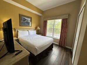 A hotel room with a bed, a television on a stand, a painting on the wall, and a window with curtains. - 2201 Spacious 3/2 Resort Condo Near Disney (Orlando)