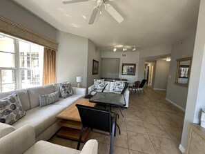 A spacious living area with a sofa, coffee table, and dining table. There is a ceiling fan, a window with curtains, and a hallway leading to another room. - 2202 Spacious 3/2 Resort Condo Near Disney (Orlando)