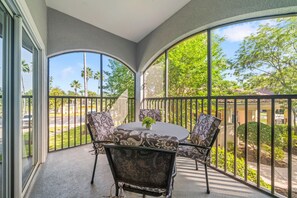 Un balcon avec une table ronde et deux chaises, un pot de fleurs et une vue sur un quartier résidentiel bordé de palmiers.
