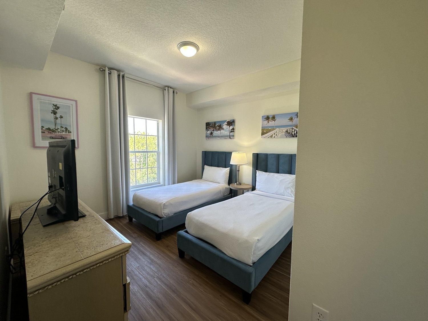 Een hotelkamer met twee bedden, een bureau met een computer, een raam met gordijnen en wanddecoratie.