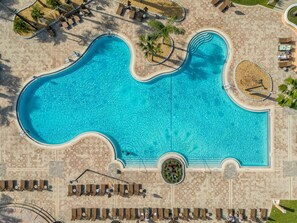 Eine Vogelperspektive auf einen Swimmingpool mit einem ungewöhnlichen Wellenmuster, umgeben von Liegestühlen und Sonnenbetten, sowie einem kreisförmigen Garten.