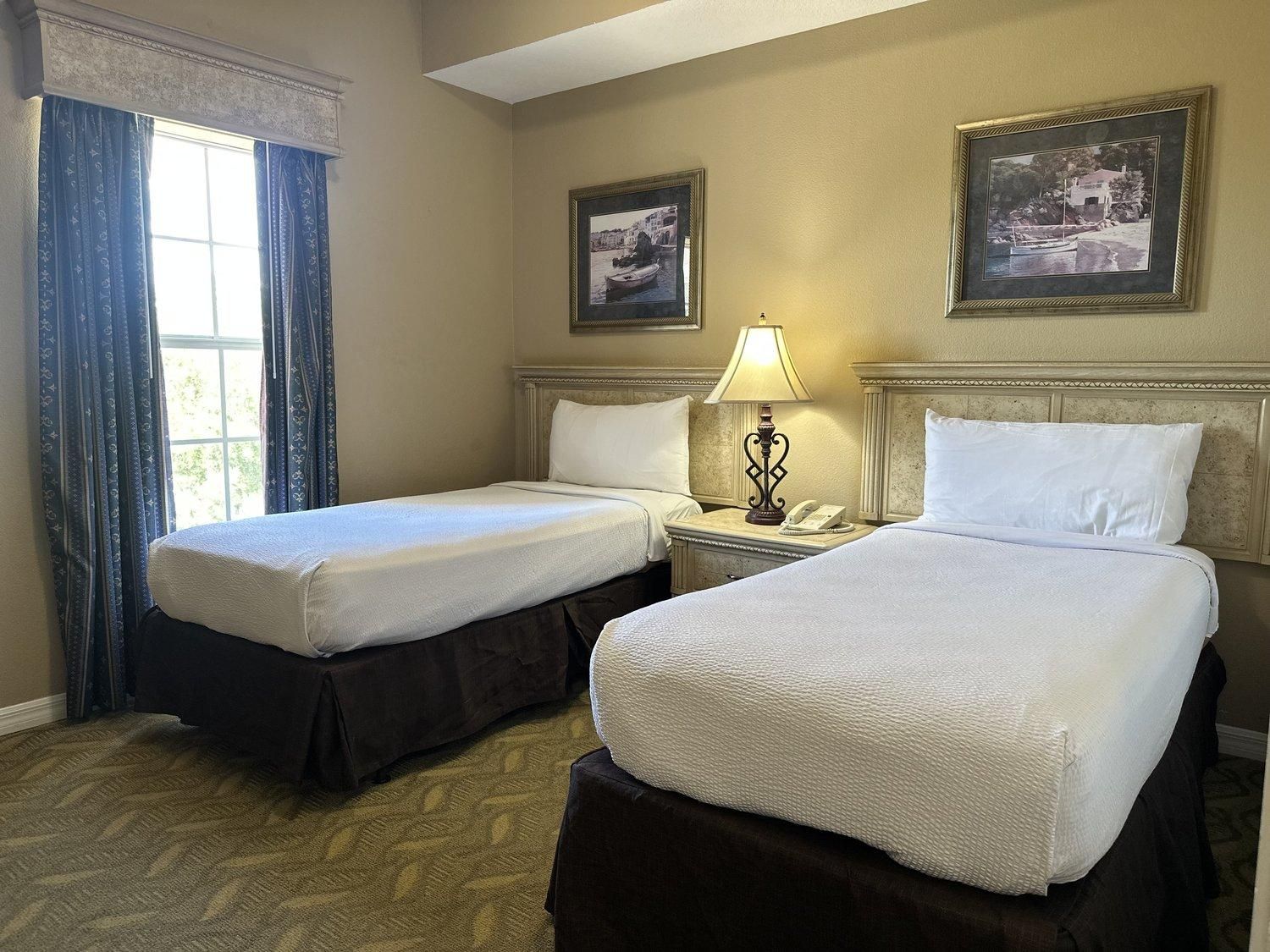 A hotel room with two single beds, a nightstand with a lamp, and two framed pictures on the wall.
