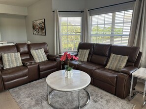 Ein Wohnzimmer mit einem braunen Ledersofa, einem runden Marmortisch und Blick auf Grünflächen durch die Fenster.