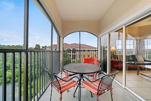 Een balkon met een tafel en stoelen, een glazen deur naar een zithoek binnen en uitzicht op gebouwen en groen.
