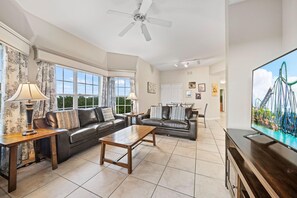 Ein geräumiges Wohnzimmer mit Sofa, Couchtisch und Fernseher. Es gibt Fenster mit Vorhängen, einen Deckenventilator und eine Tür zu einem anderen Raum.