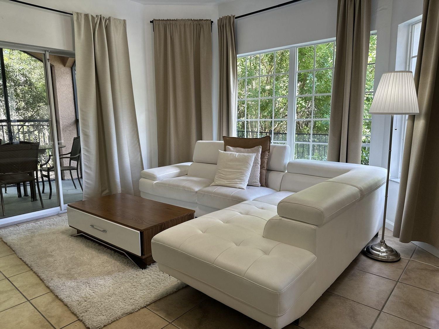 Ein modernes Wohnzimmer mit einem weißen Sofa, einem Holztisch und Blick auf Grünanlagen durch große Fenster.