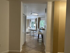 A hallway leading to a dining area with a table and chairs, a window with curtains, and a ceiling light fixture.