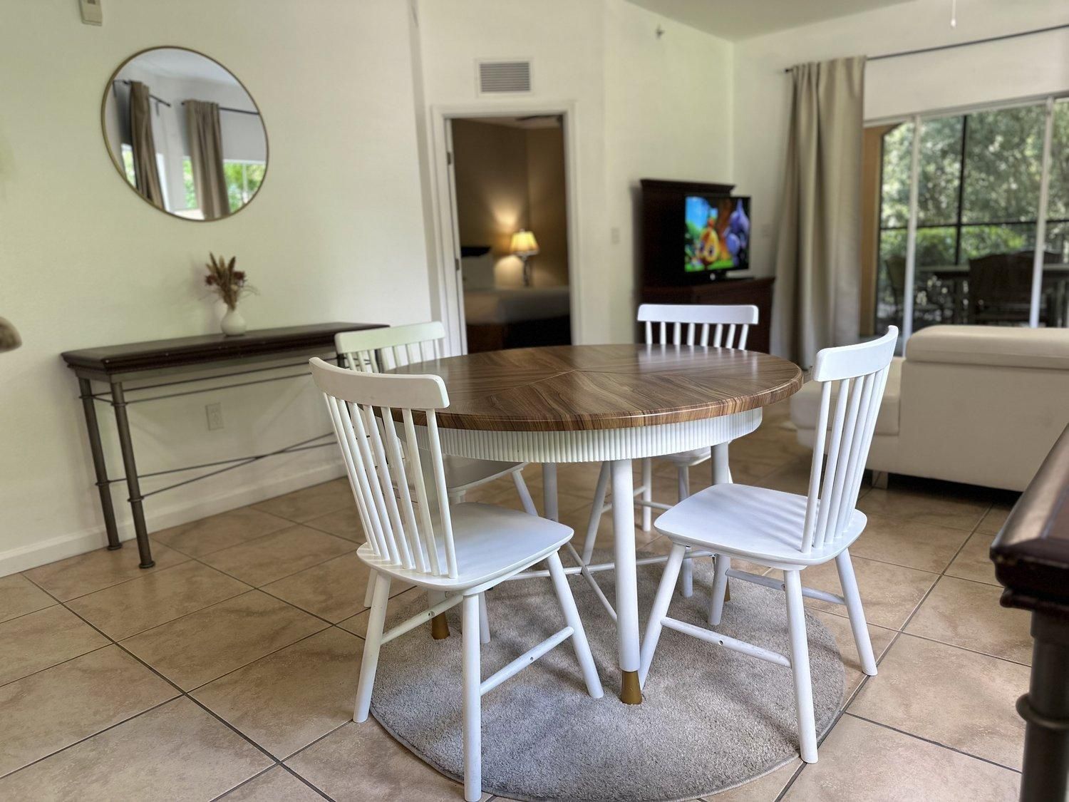 Un coin repas avec une table ronde en bois et des chaises blanches, une petite console, un miroir et un téléviseur en arrière-plan.