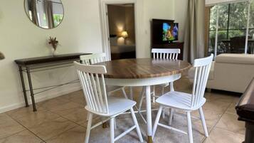 Un coin repas avec une table ronde en bois et des chaises blanches, une petite console, un miroir et un téléviseur en arrière-plan.