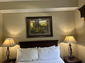 A hotel room with a bed, two bedside lamps, a framed picture on the wall, and a wooden headboard.
