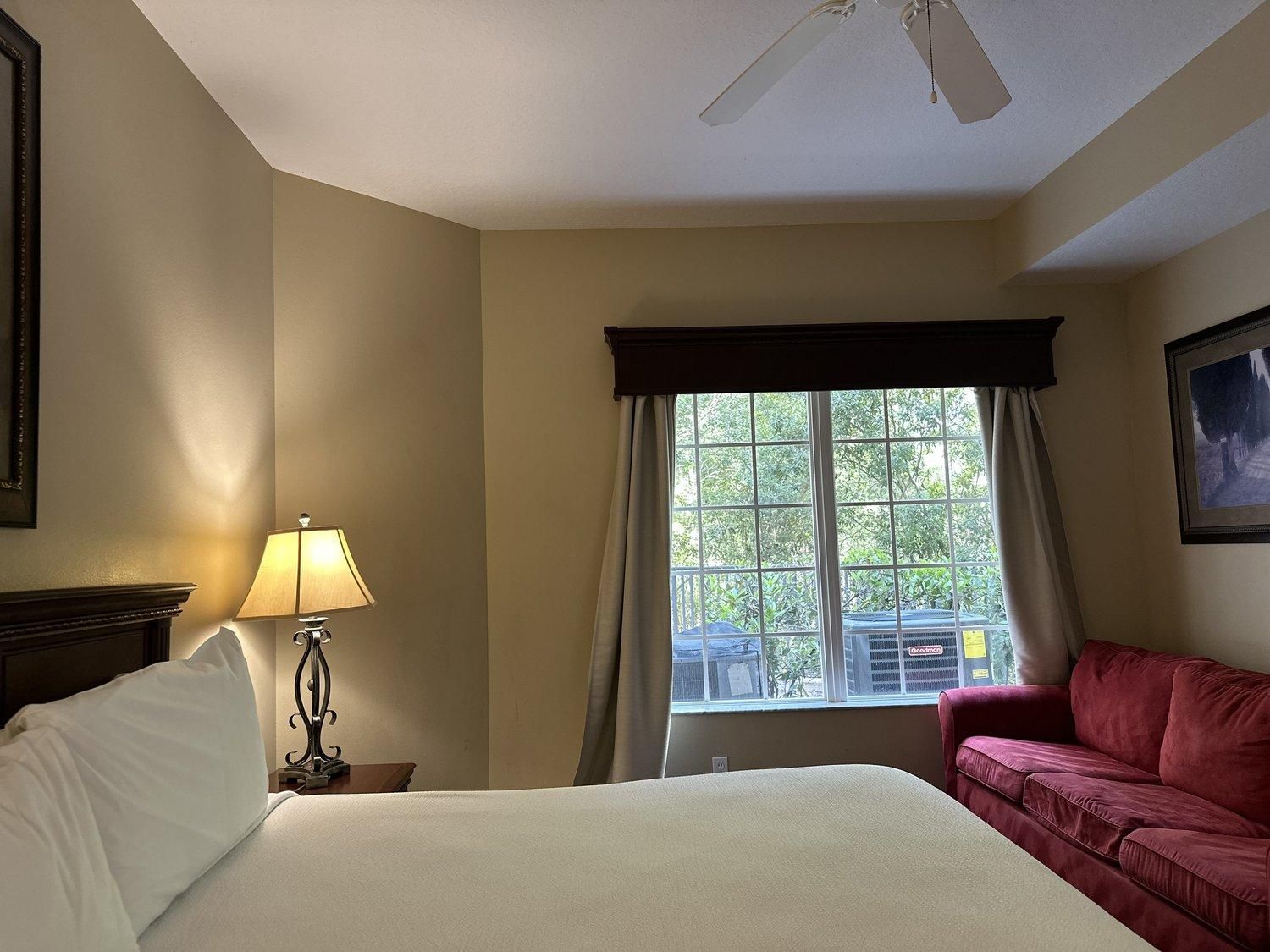 A hotel room with a bed, a red sofa, a window with curtains, a lamp, and a ceiling fan.