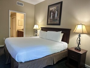 A hotel room with a large bed, a nightstand with a lamp, a framed picture on the wall, and a door leading to a bathroom.