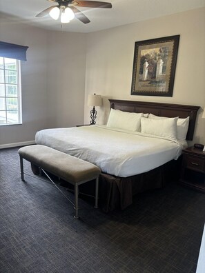 Une chambre avec un grand lit, un banc, une table de chevet, un ventilateur de plafond, une fenêtre avec des stores et un tableau encadré au mur.