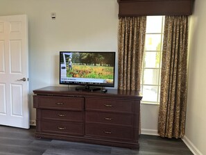 Un téléviseur posé sur un meuble en bois, une porte blanche et une fenêtre ornée de rideaux à motifs.